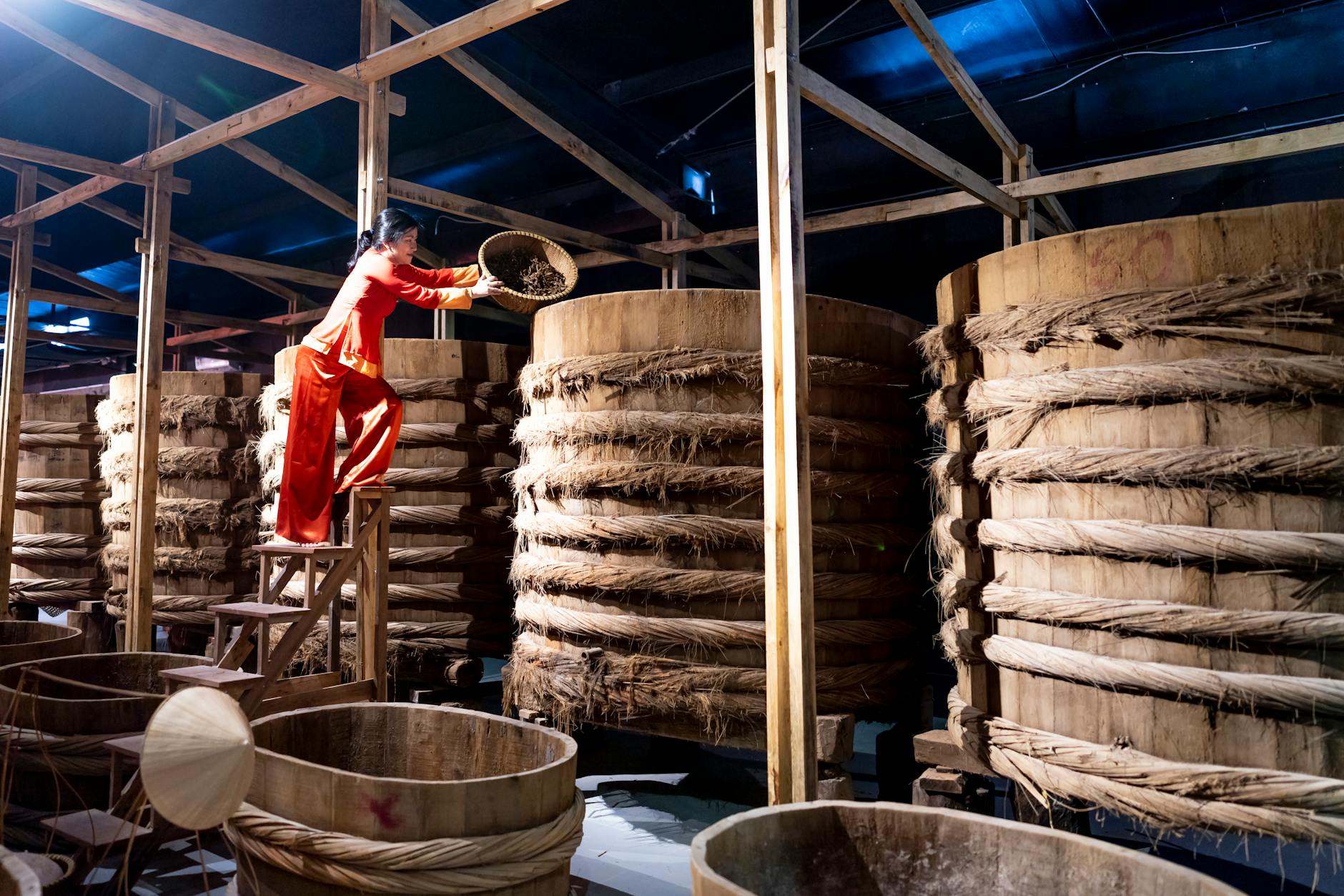 Full body of female adding ingredient from bowl into wooden barrel with fish sauce in factory
