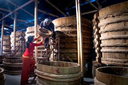 Woman helping male colleague standing on ladder while working in fish sauce factory together