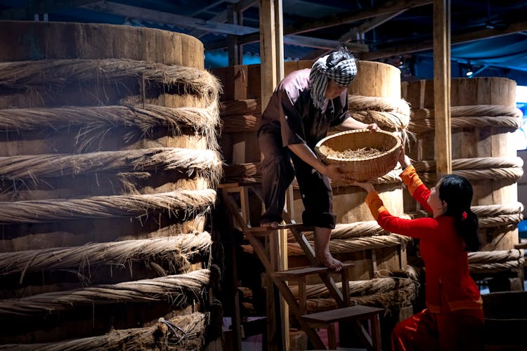 Asian Man Giving Bowl To Ethnic Colleague In Barrel Storage