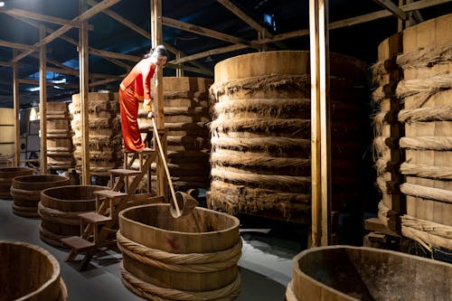 Full body of Asian female worker with instrument standing near row of barrels in fish sauce factory