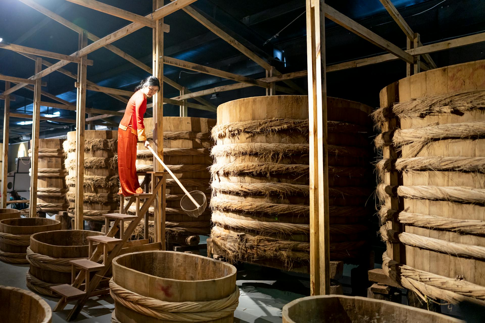 Woman in red clothing overseeing fermentation process in a traditional wooden barrel warehouse.