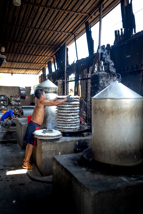 Free Full length of man standing near metal barrels and machines while making noodle on caps in factory in Vietnam Stock Photo