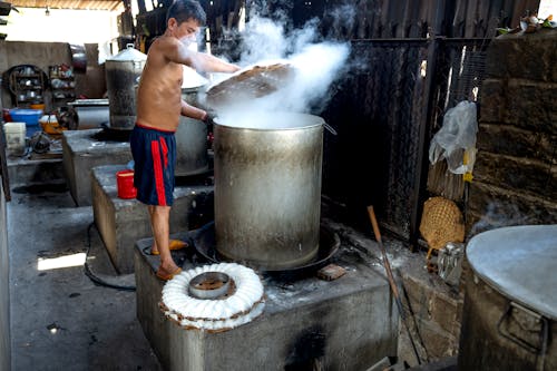 Full body side view of guy standing near metal barrels and machines while making noodle on caps in factory in Vietnam