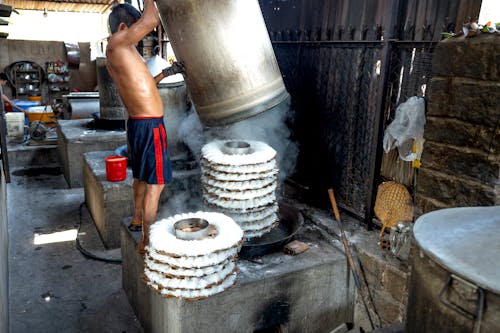 Side view of anonymous male in shorts while making noodle on caps near metal barrels and machines on manufacture in Vietnam