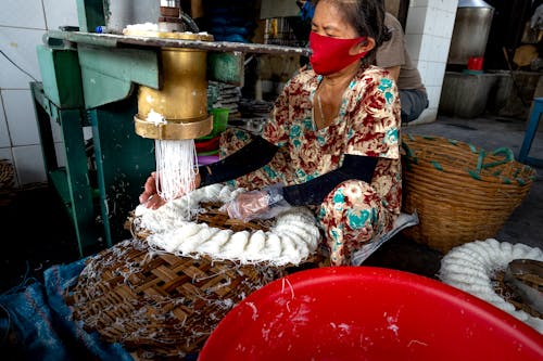 Woman in casual outfit and mask making noodle on wooden caps on metal machine in factory in Vietnam