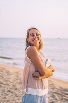 A smiling woman holding a notebook, enjoying a serene sunset by the sea.
