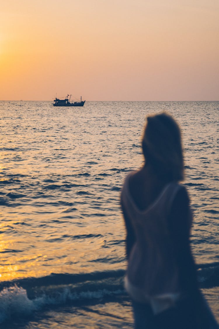 A Woman Standing On The Shore While Looking At The Boat Sailing During Sunset