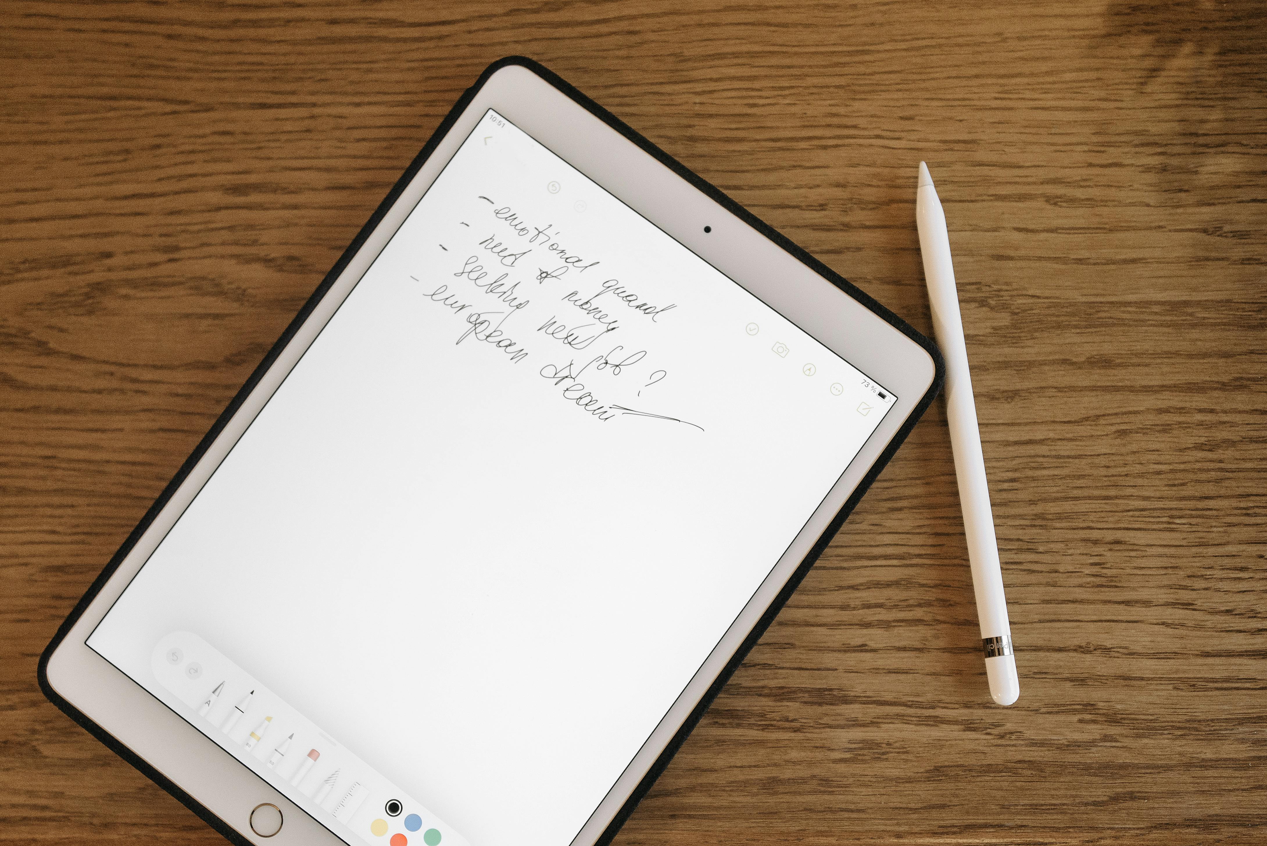 Flat lay of a tablet with handwritten notes and a stylus on a wooden desk background.