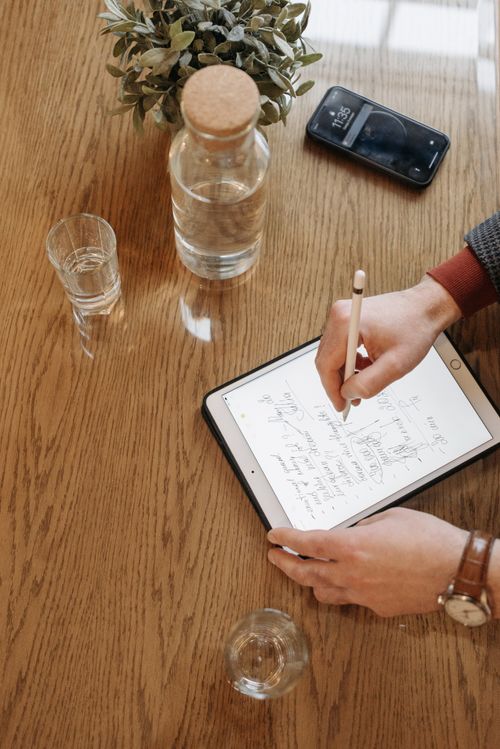 Free Individual writing with Apple Pencil on an iPad, surrounded by everyday items on a wooden table. Stock Photo