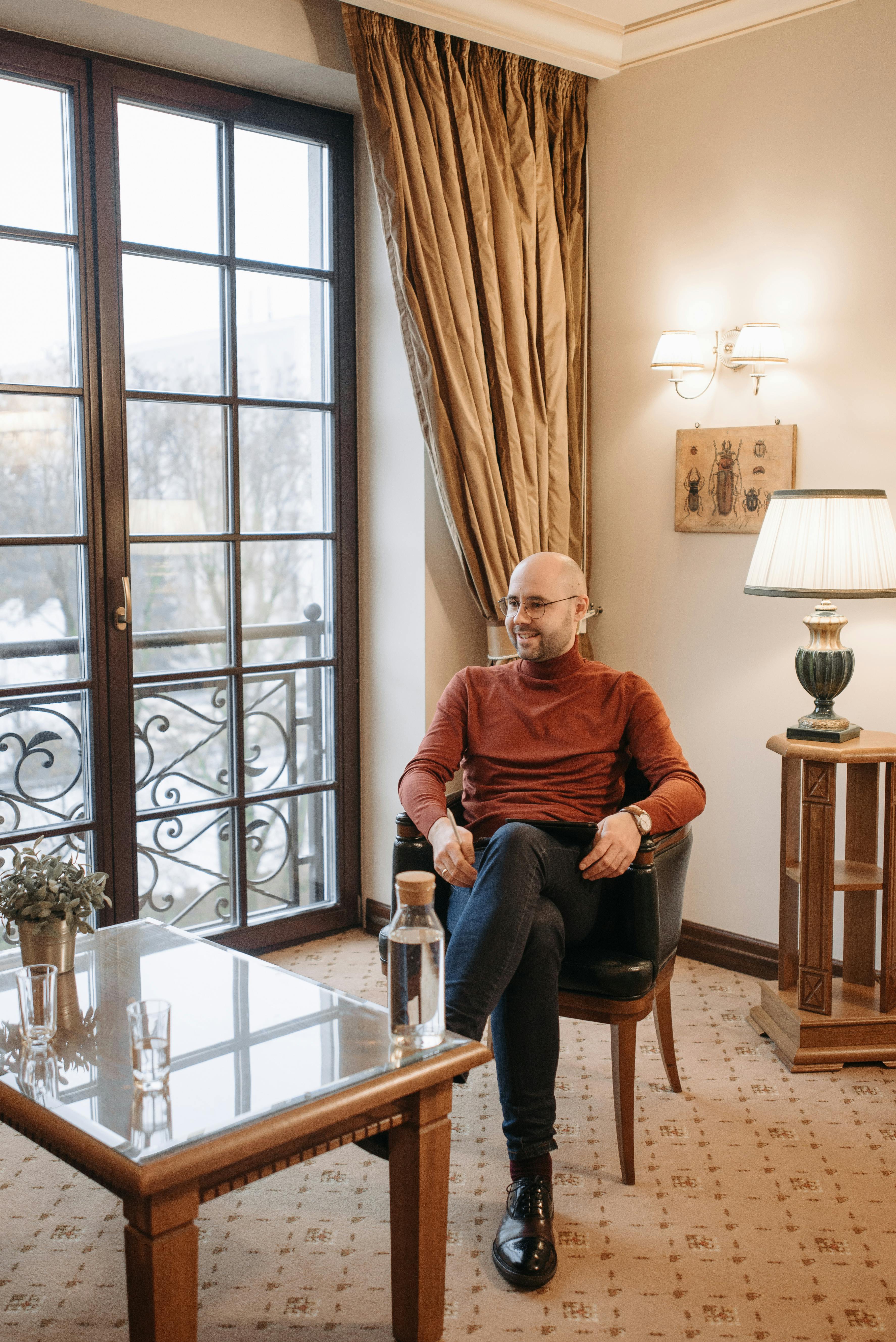 Bald man sitting in a cozy room by glass doors, wearing a turtle neck and smiling.