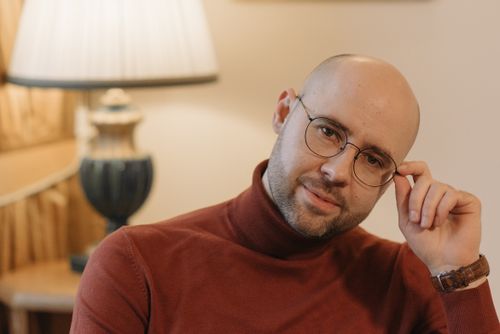 Free Bald man smiling in a brown turtleneck, adjusting glasses in cozy indoor setting. Stock Photo