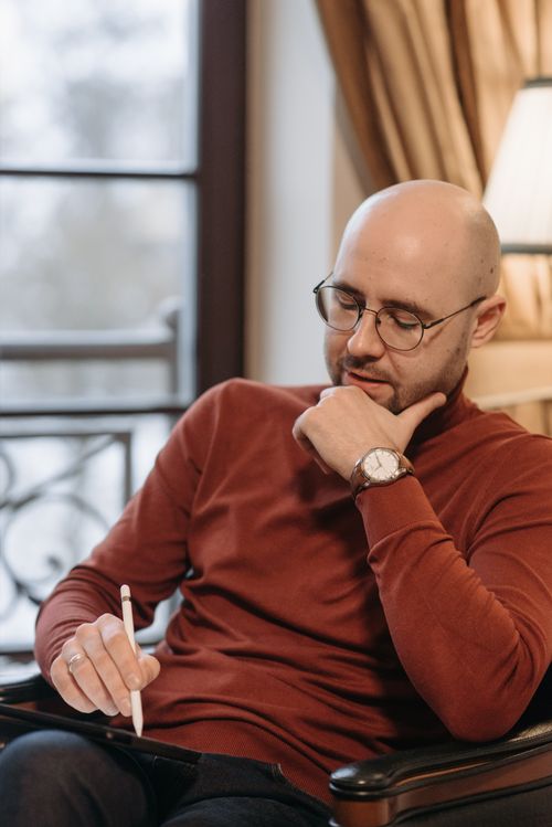 Free Bald man with eyeglasses sitting indoors, writing thoughtfully with a pen. Stock Photo