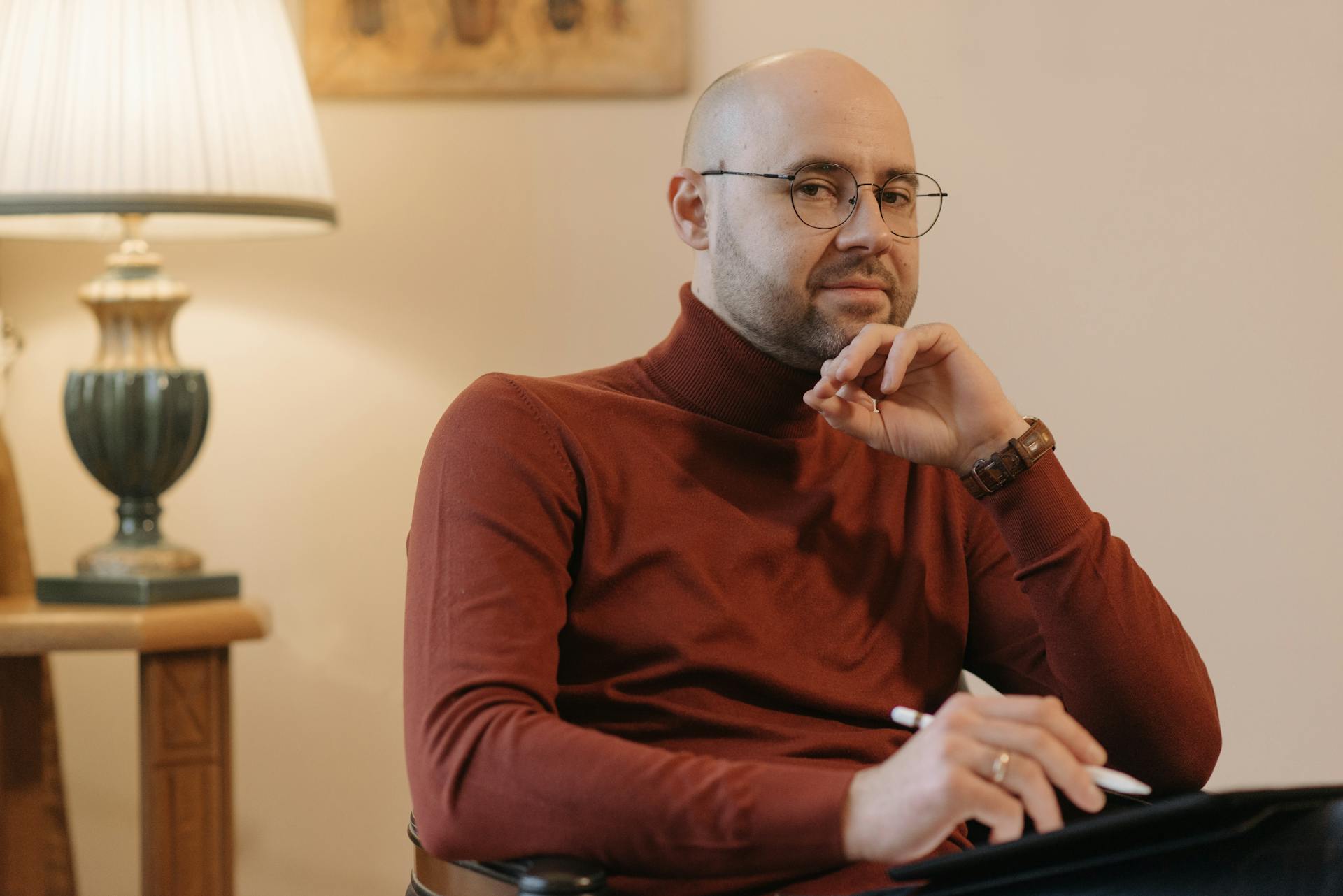 A bald man with eyeglasses holding a pen, sitting thoughtfully indoors wearing a red turtleneck.