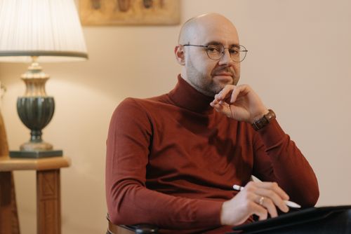 Free A bald man with eyeglasses holding a pen, sitting thoughtfully indoors wearing a red turtleneck. Stock Photo