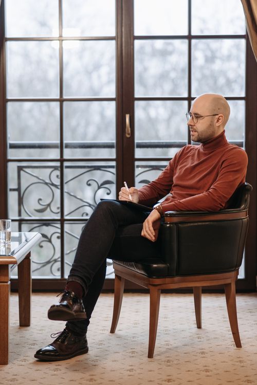 Free Bald man in sweater sitting with notes near large glass door, looking thoughtful. Stock Photo