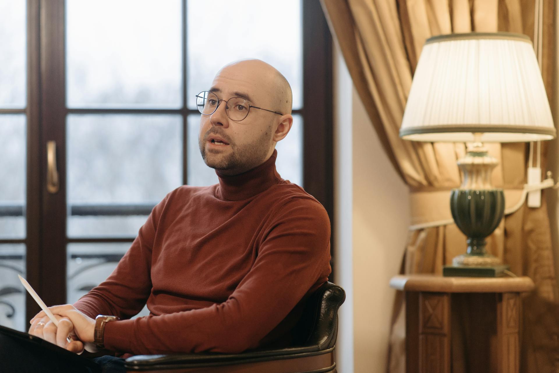 Bald man in glasses speaking while seated indoors, with a warm lamp and curtains in the background.