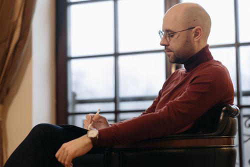 Free A bald, bearded man in a red sweater writing in a notebook indoors. Stock Photo