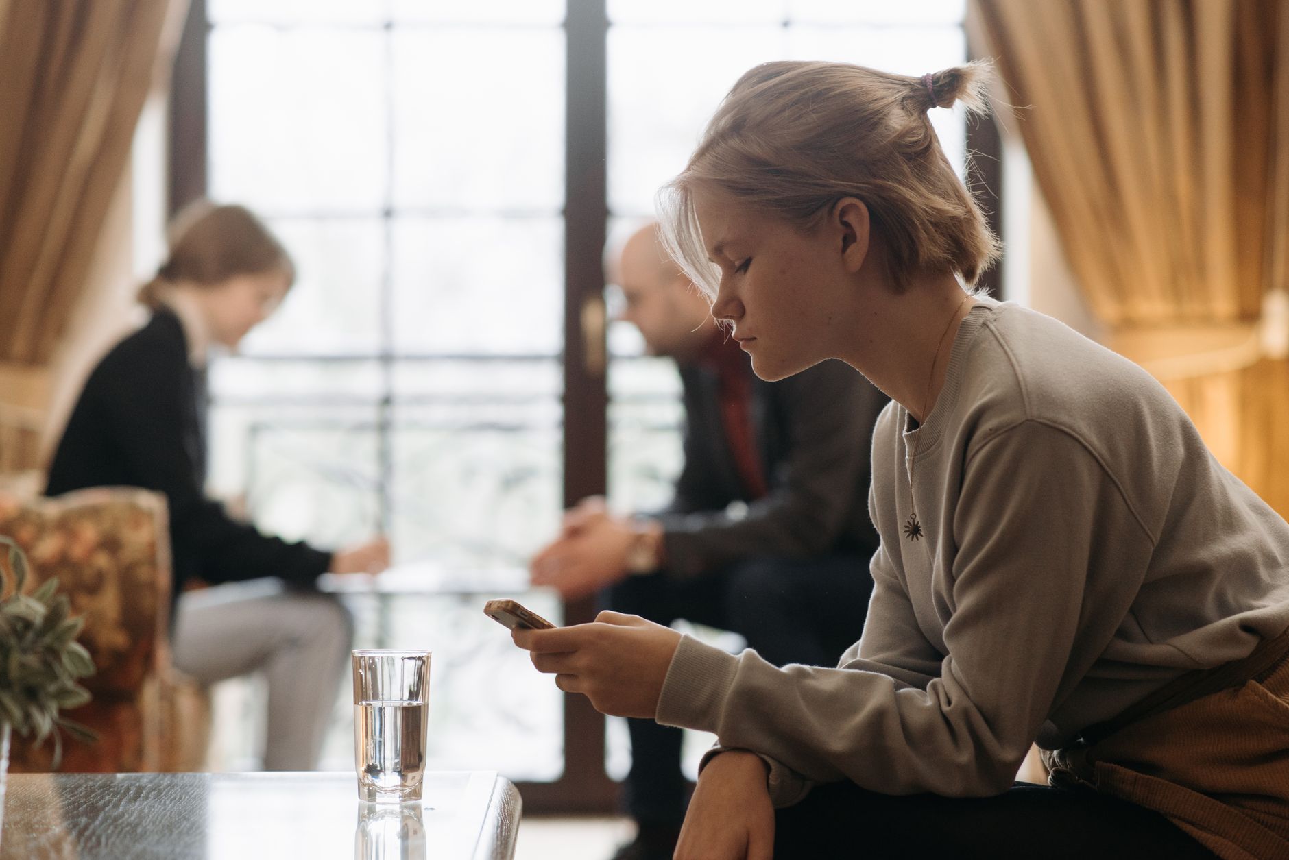 Teenager focused on smartphone in cozy indoor setting with blurred adults in background.
