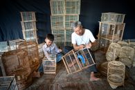 Aged ethnic craftsman with gray hair creating handmade souvenir bird cages while little grandson helping and picking wooden bamboo sticks