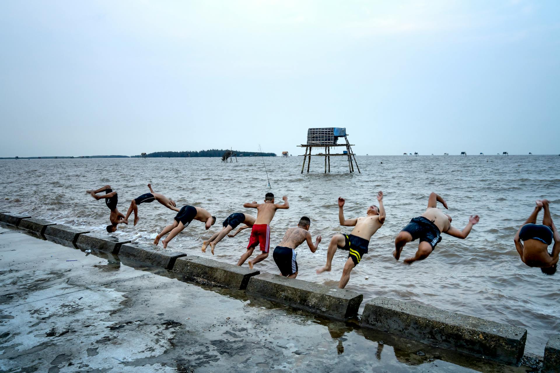 Anonymous men in swimming trunks performing tricks in air while jumping into ocean against lifeguard tower under light sky