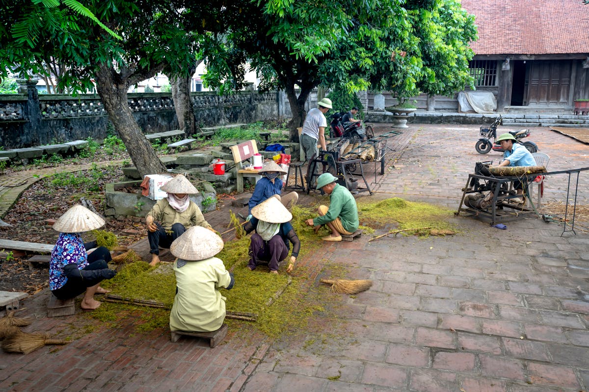Unrecognizable ethnic people in conical hats placing ground tea foliage on tray while sitting on walkway in town