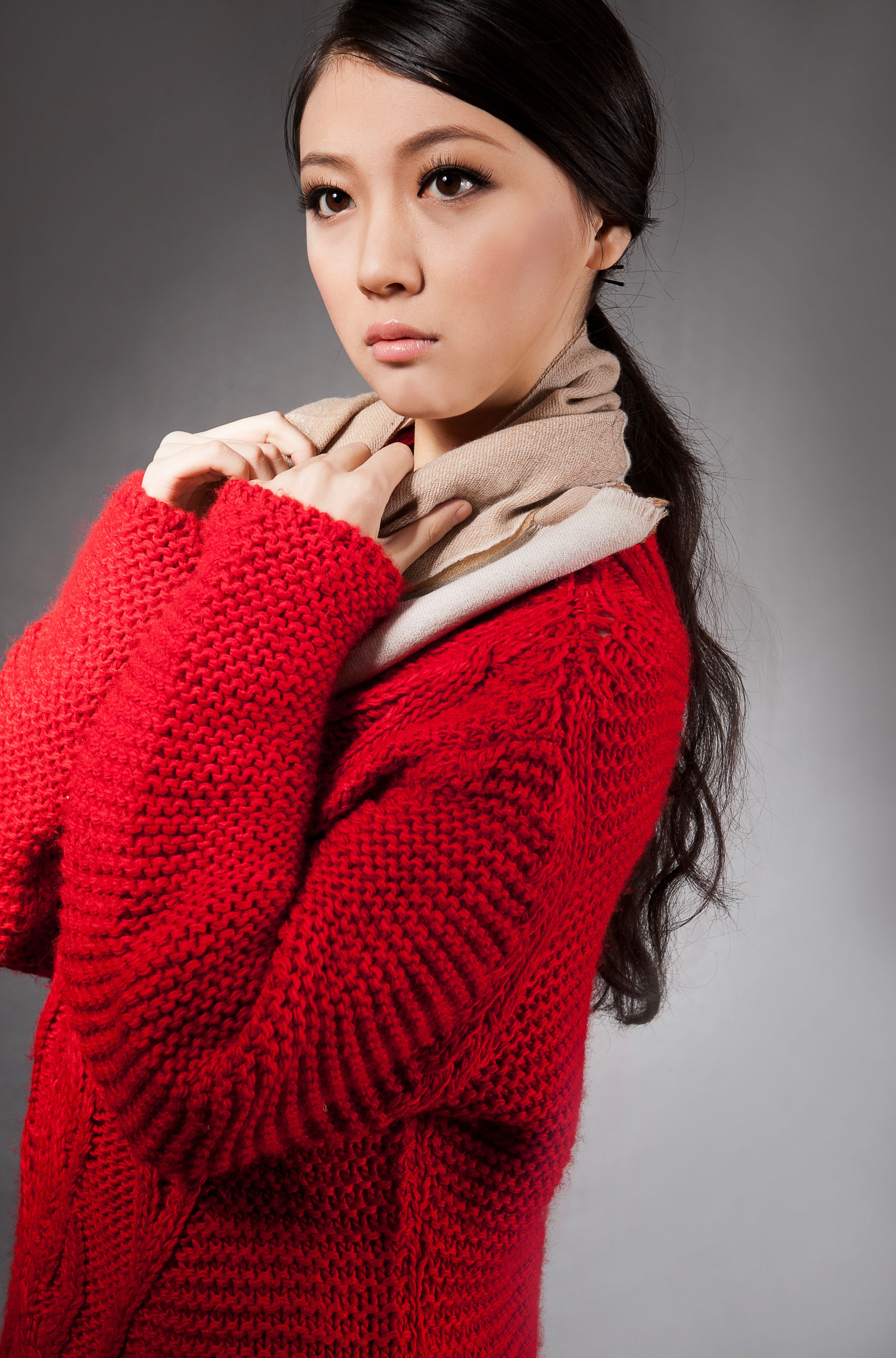 Side view of crop pensive young Asian female model with long dark hair in fashionable knitted cardigan and scarf standing against gray background and looking away