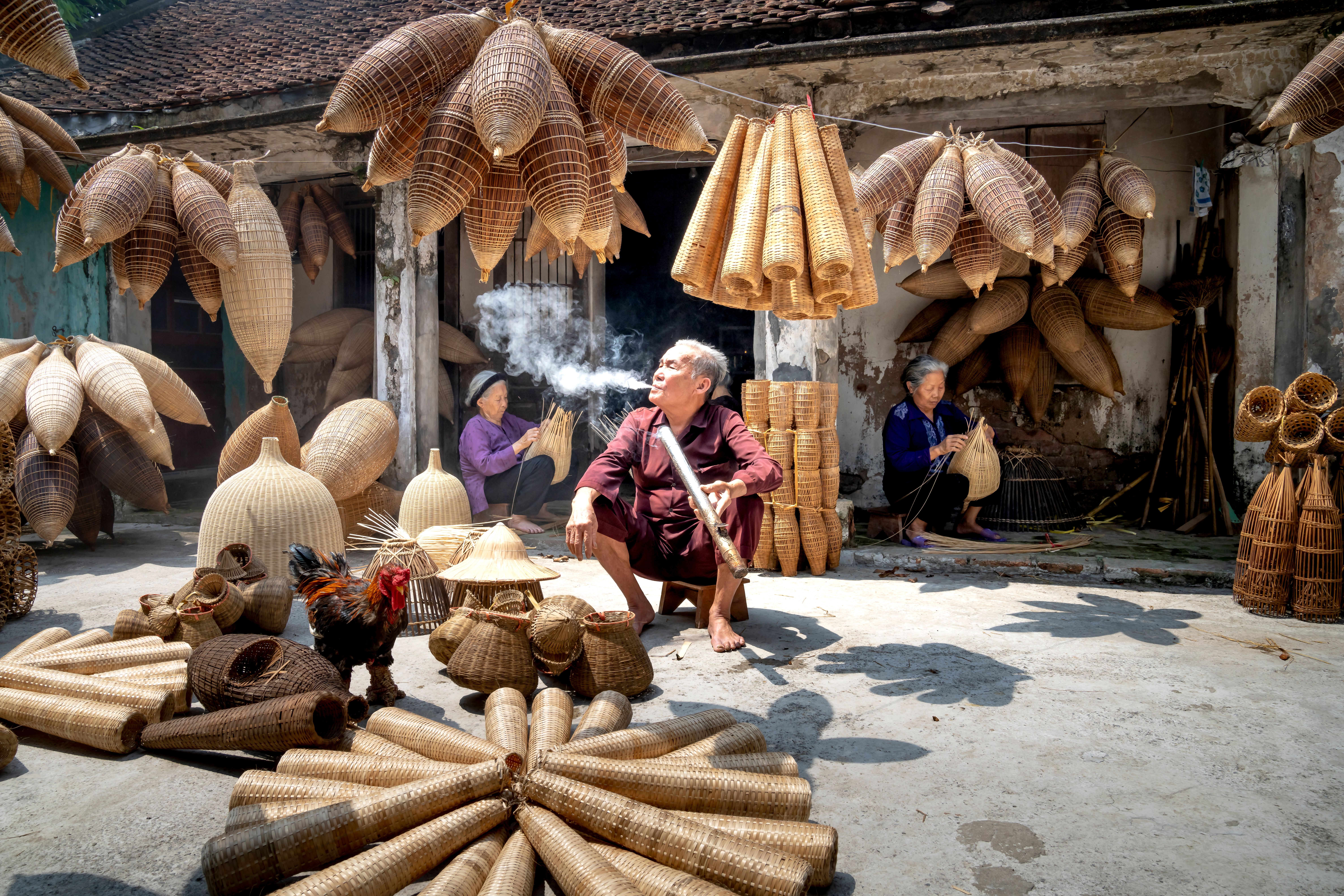 Relaxing ethnic elderly male smoking near aged women making timber traditional Vietnamese fish traps in rustic yard