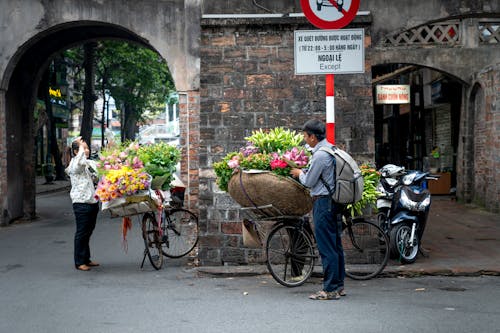 Free Side view of male in casual clothes standing near bicycle with baskets of colorful flowers of vendor and buying flowers from woman on town street Stock Photo