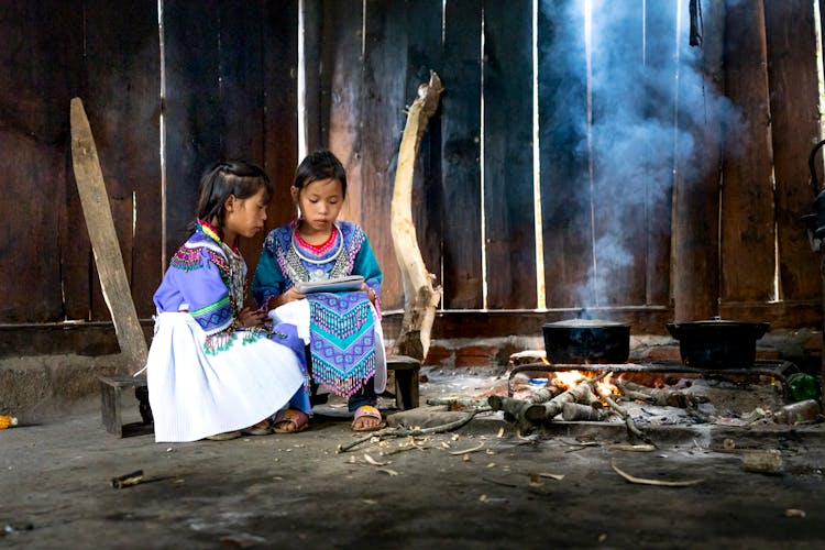 Little Girls In Traditional Clothes Reading By Fire In Hut