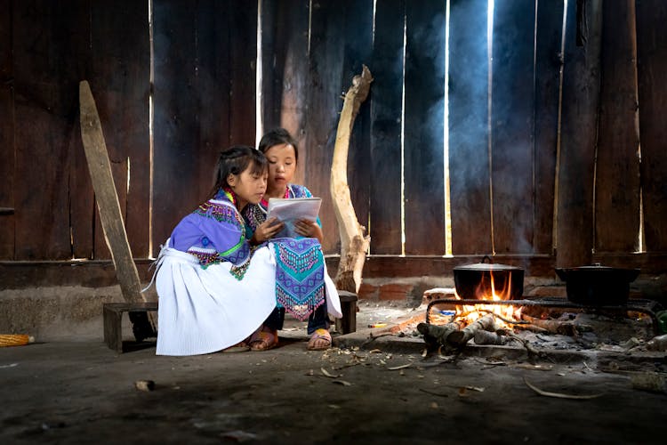 Girls In Traditional Dresses Sitting And Reading By A Fire In A Wooden Shed