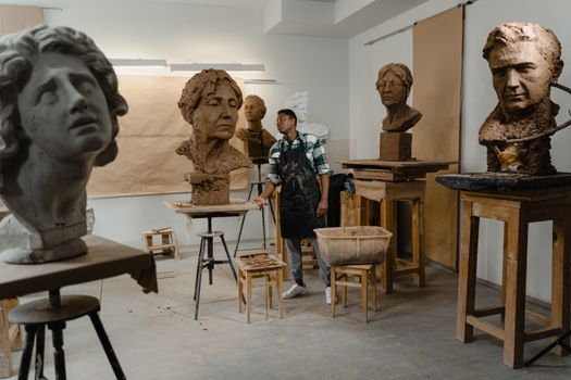 A sculptor working on clay busts in an art studio, surrounded by handmade sculptures.