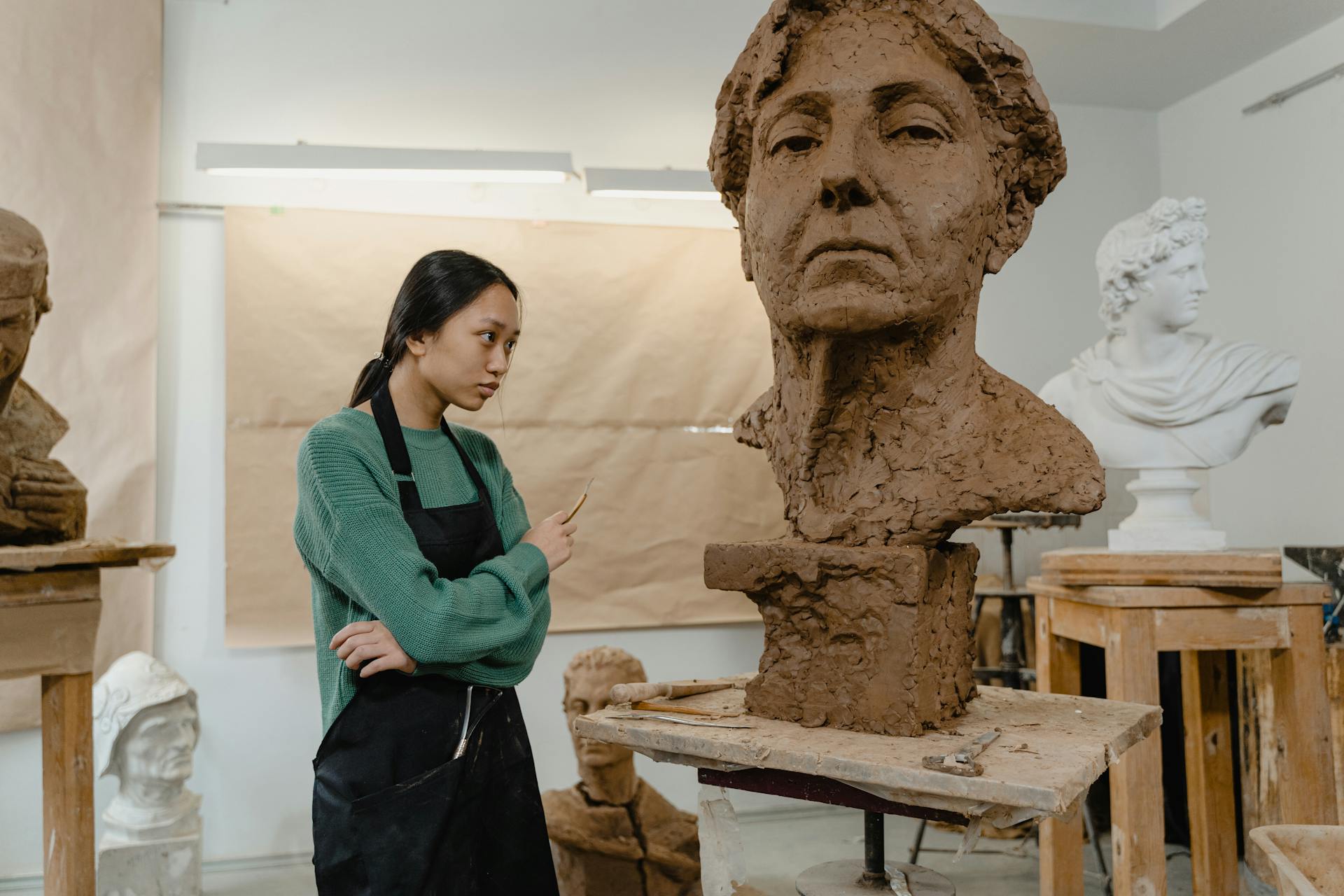 A young woman sculptor creating a clay bust in an art studio, surrounded by sculptures.