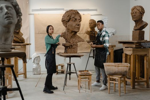 Two artists work on clay sculptures in a well-lit art studio.