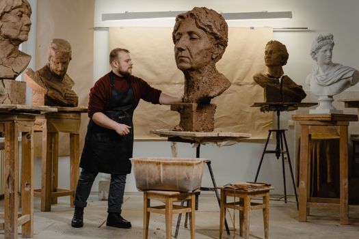 A sculptor in an art studio creating detailed clay busts, surrounded by various sculptures.