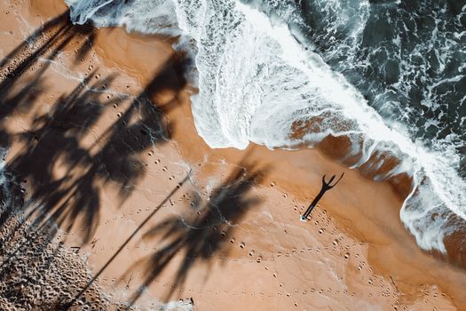 An aerial view of a beach with palm shadows and a lone figure enjoying the summer waves.