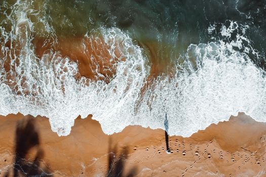 Drone view of waves meeting sandy shore, capturing footprints and surf in a vibrant summer setting.