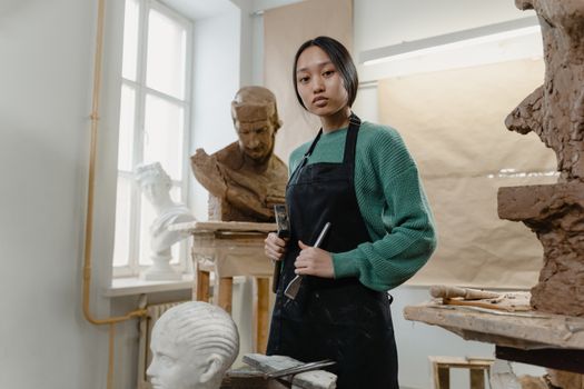 A female sculptor working in an art studio surrounded by clay sculptures and tools.