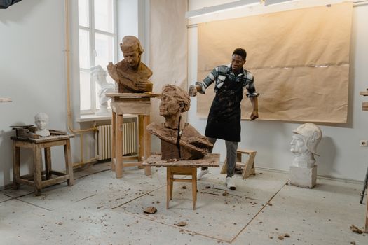 Artist in art studio sculpting clay with hammer, surrounded by sculptures. Creative workspace vibe.