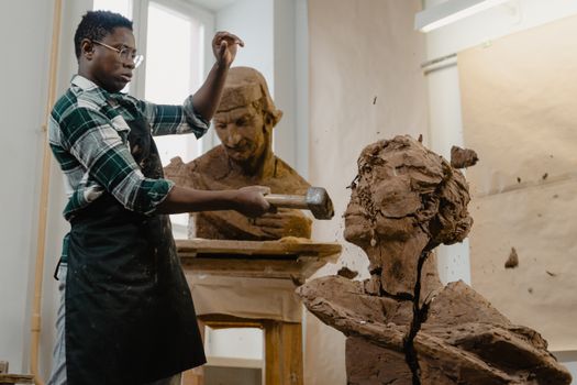 A man wearing an apron skillfully sculpts a clay bust in an art studio using a hammer.