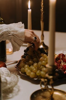 A hand reaches for grapes at a candle-lit dinner table set with elegant dishes and brass candlesticks.