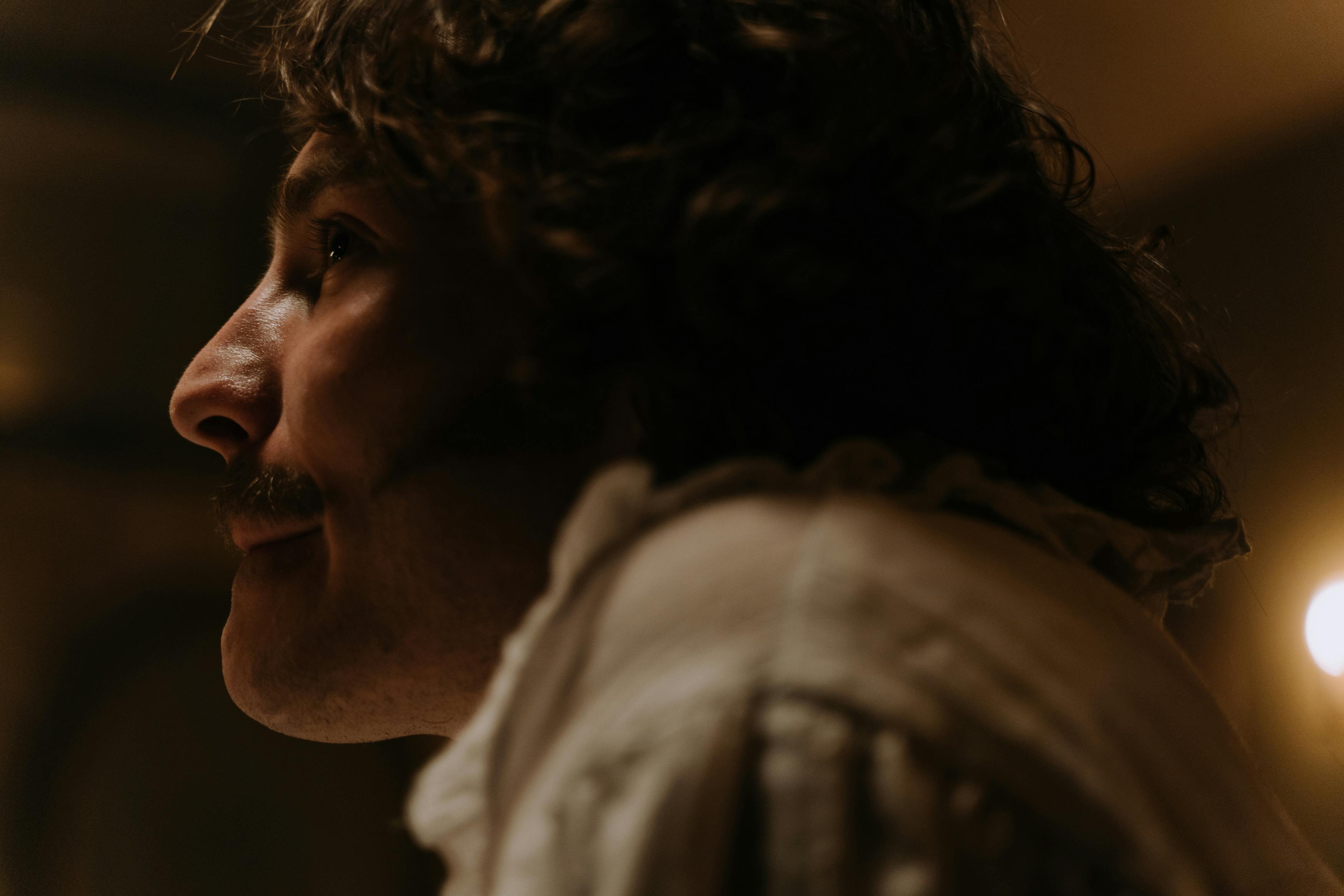 Side profile of a man with curly hair and mustache in dim lighting, evoking a vintage feel.
