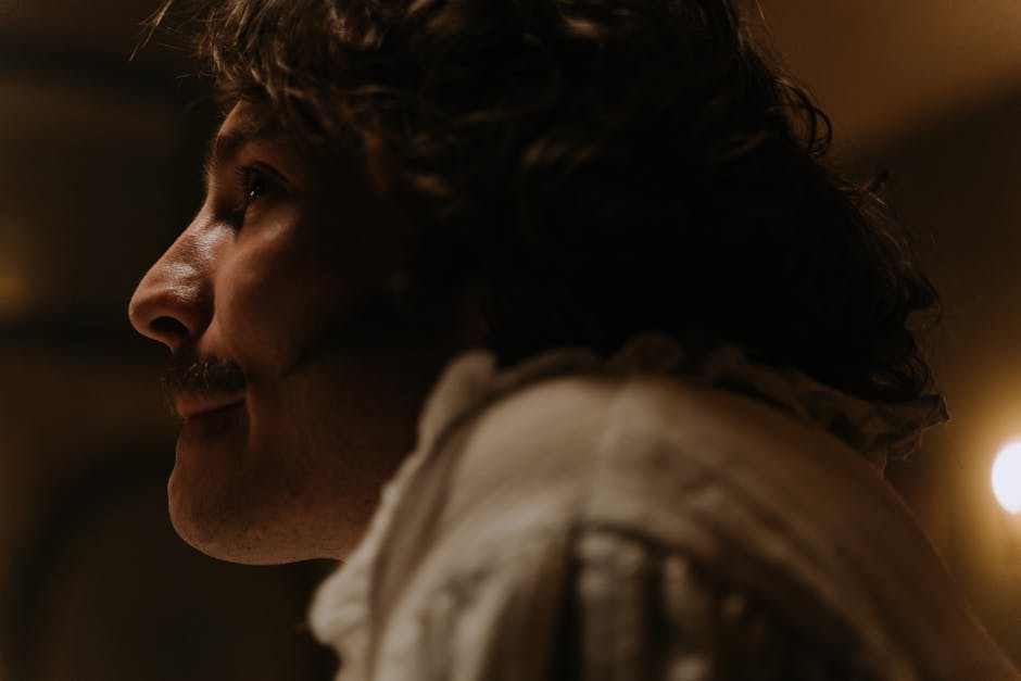 Side profile of a man with curly hair and mustache in dim lighting, evoking a vintage feel.