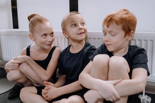 Three children sit joyfully on a floor indoors, dressed in black dance clothing.