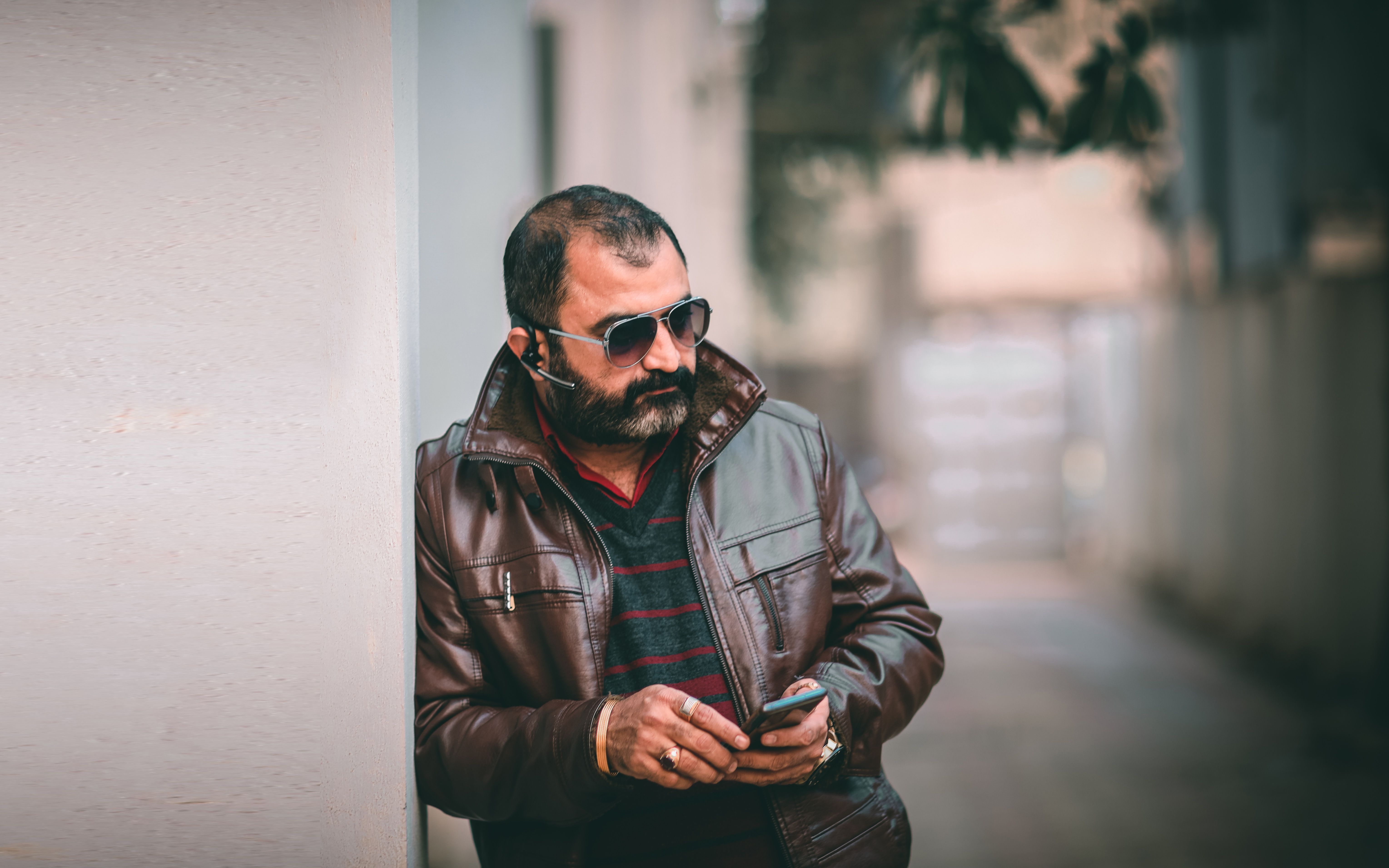 A bearded man wearing a leather jacket and sunglasses using a smartphone in an urban setting.