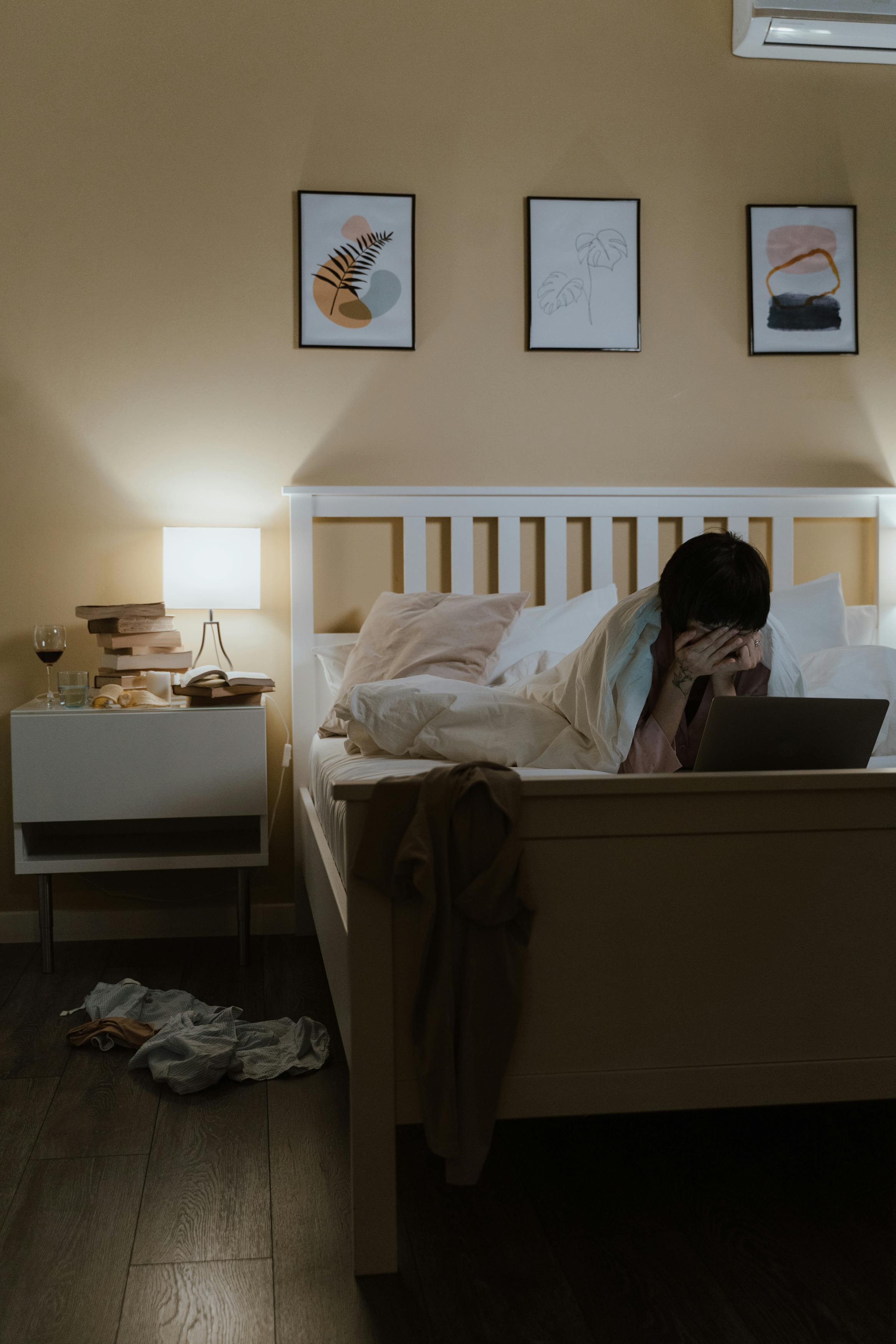 Woman sitting in bed with laptop, looking sad in dimly lit bedroom.
