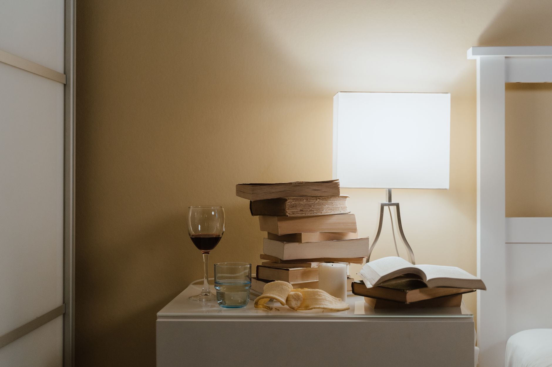 A minimalist bedroom nightstand with books, a lamp, and a glass of wine for a cozy evening vibe.