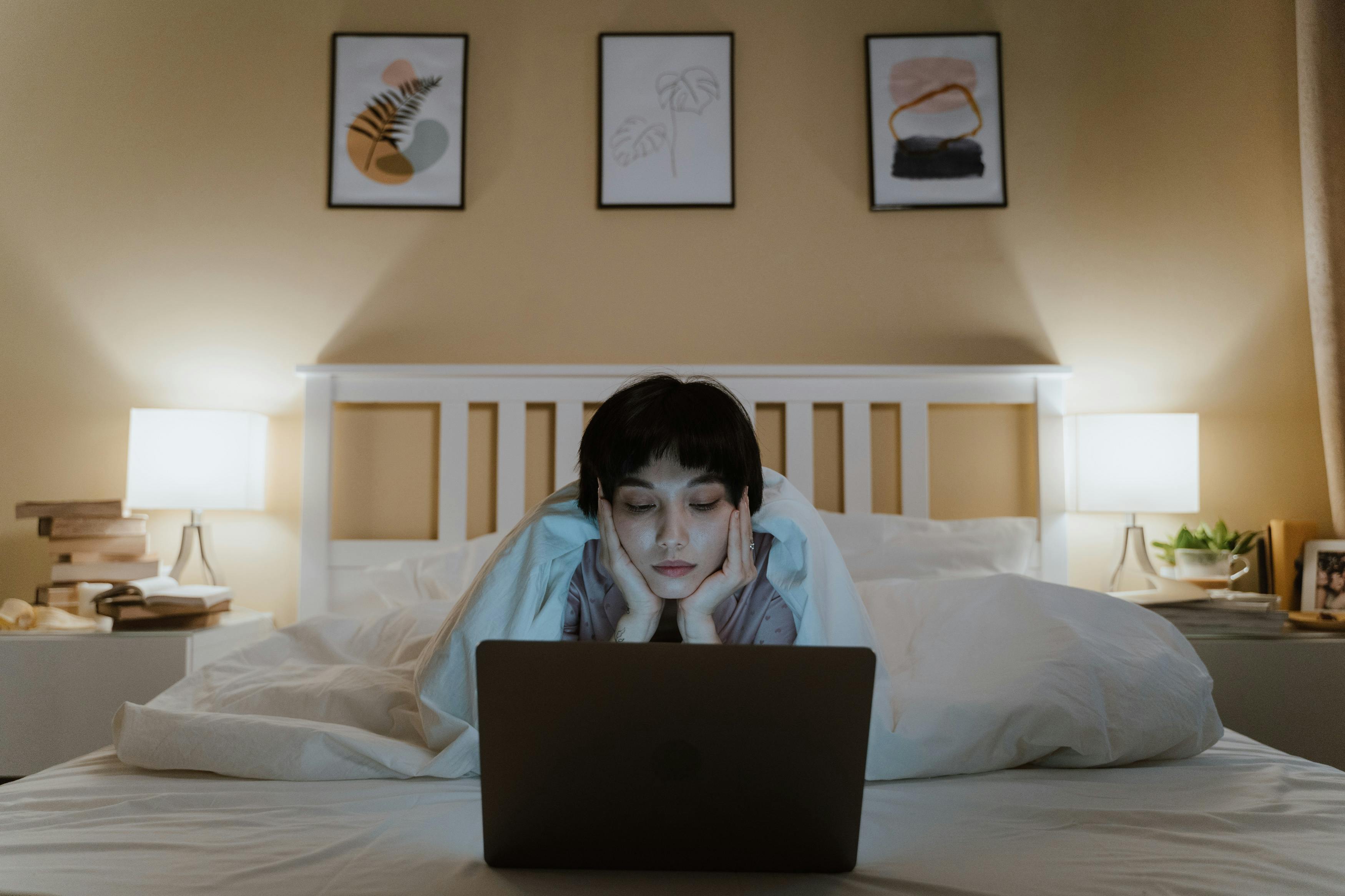 a woman under white blanket lying on the bed while looking at the screen of a laptop