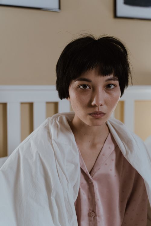 Free A woman with a short haircut wrapped in a white blanket sits on a bed, looking contemplative indoors. Stock Photo