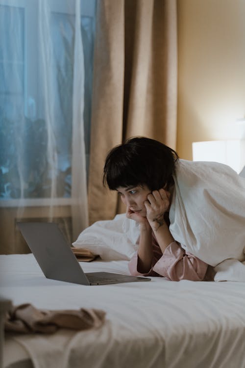 A Woman Covered with White Blanket Sitting on the Bed while Looking at the Laptop