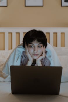 A woman comfortably browsing on her laptop while lying in bed, under a warm blanket.